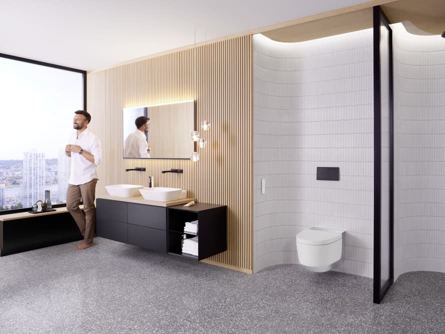 Muž ve velké koupelně s tmavým nábytkem a sprchovým WC Geberit AquaClean Muž ve velké koupelně s tmavým nábytkem a sprchovým WC Geberit AquaClean